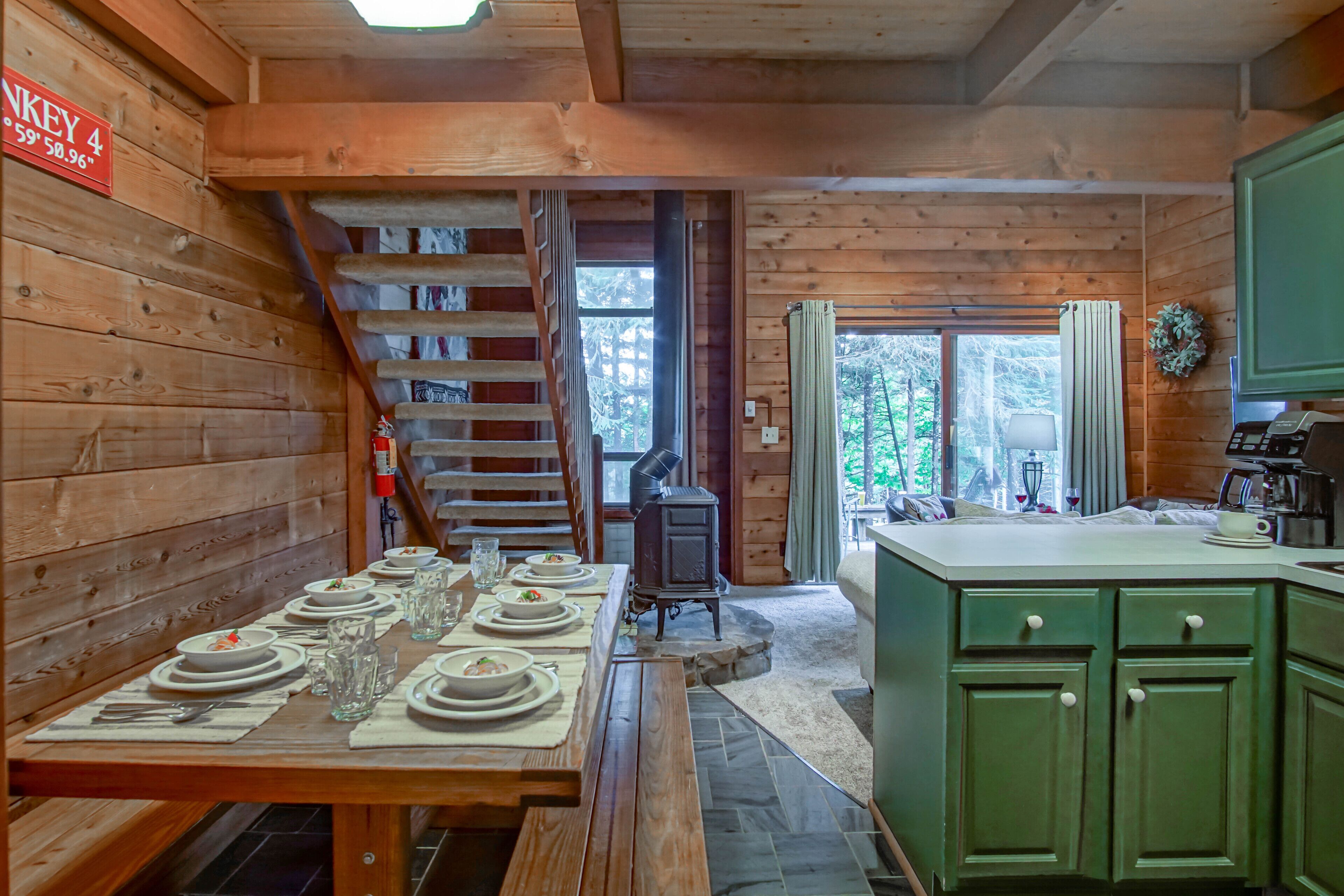 A wooden cabin interior with a dining table set for four, a wood-burning stove, and a green kitchen area.