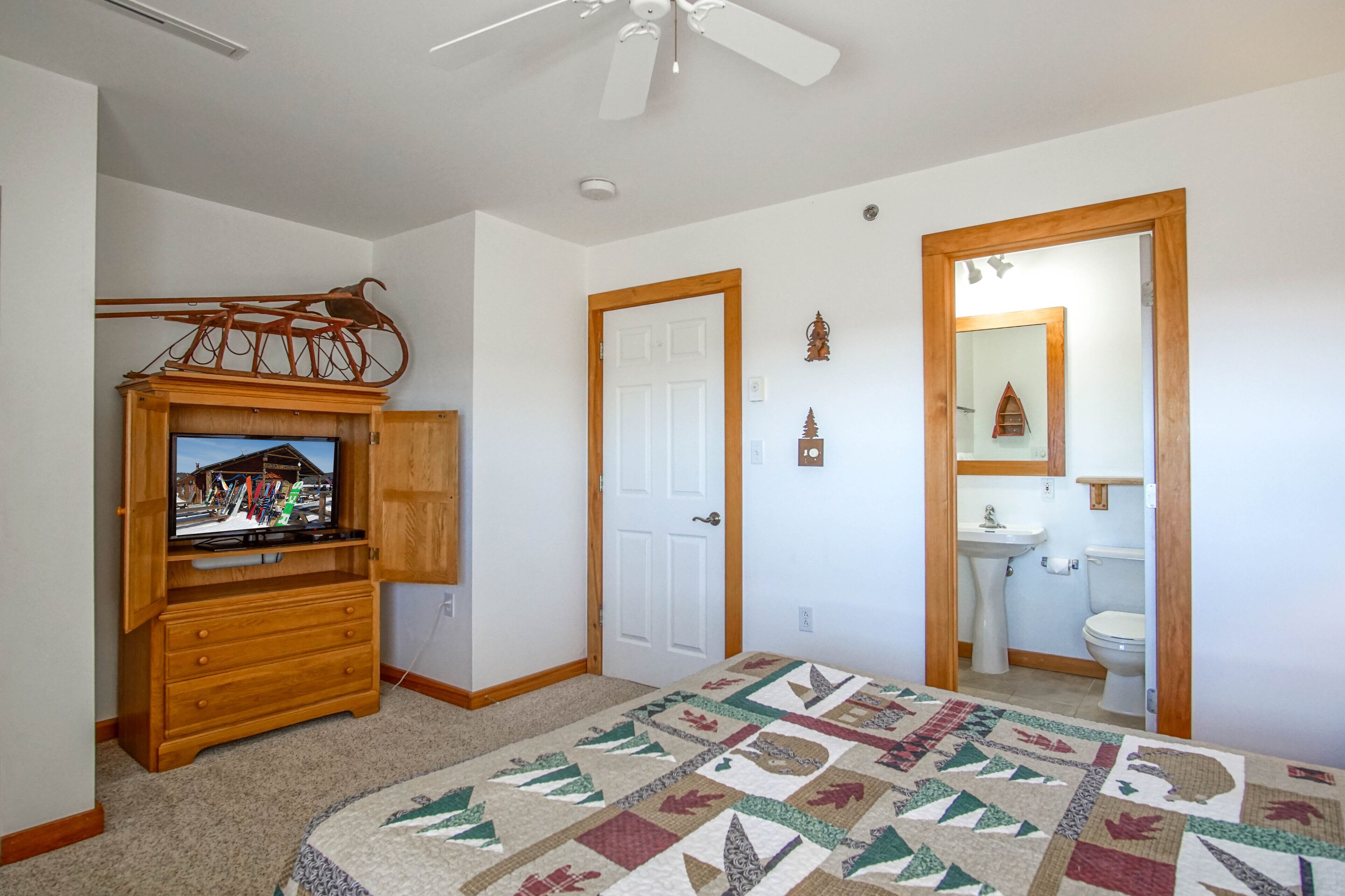A bedroom with a wooden dresser, a television on top, a ceiling fan, a door, and a bathroom with a sink and toilet.