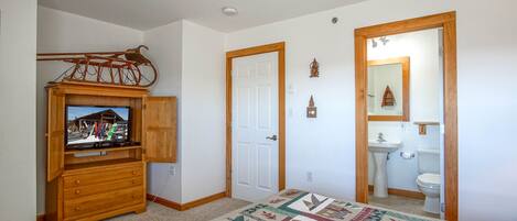 A bedroom with a wooden dresser, a television on top, a ceiling fan, a door, and a bathroom with a sink and toilet.