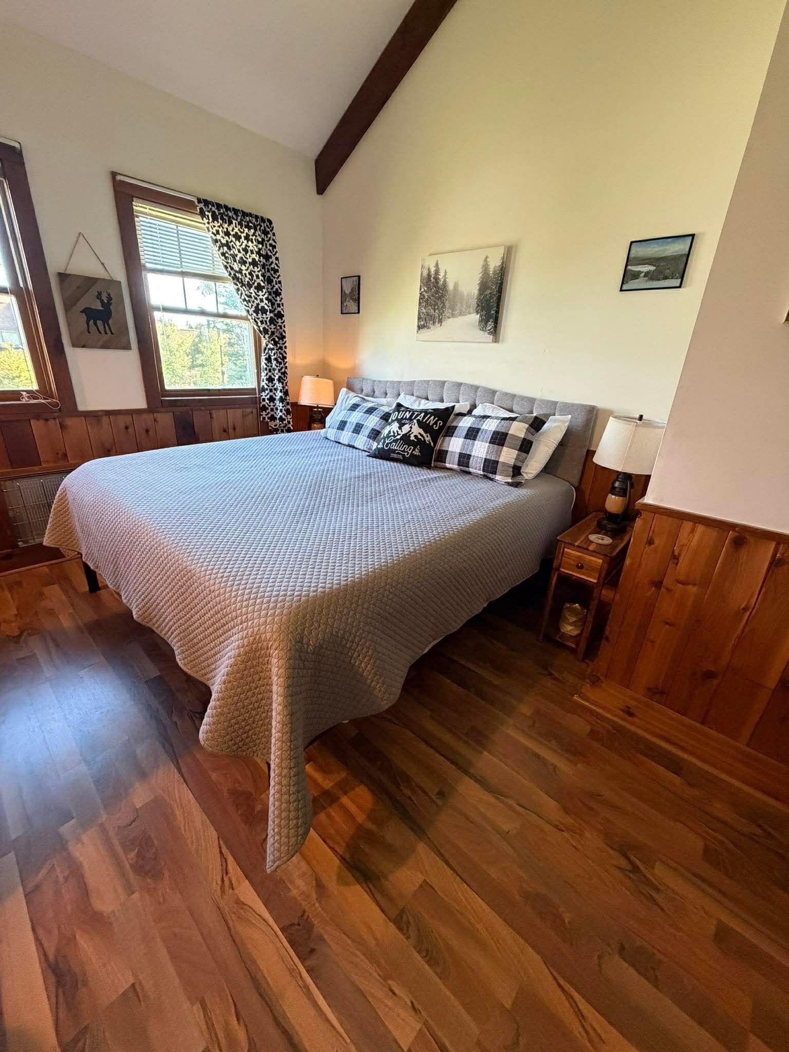 Quarto com cama grande, piso de madeira, janela com persianas, luminária e quadro na parede.