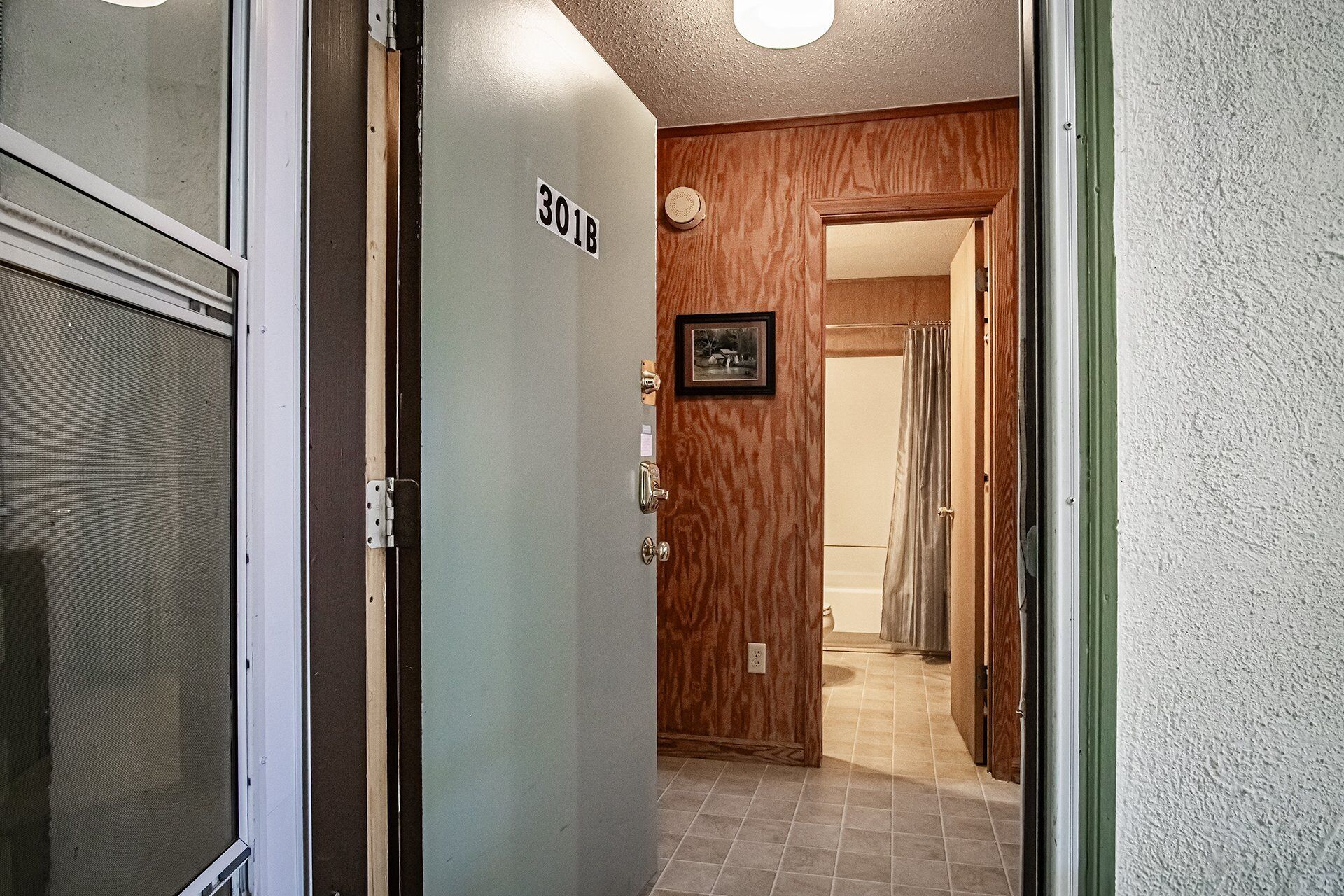 Un couloir avec une porte portant l’inscription « 301B », qui donne sur une pièce équipée d’une douche et ornée d’un tableau encadré au mur.