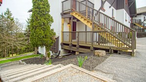 En toetages bygning med en trætrap og balkon, omgivet af en anlagt have med et lille træ og grusveje.