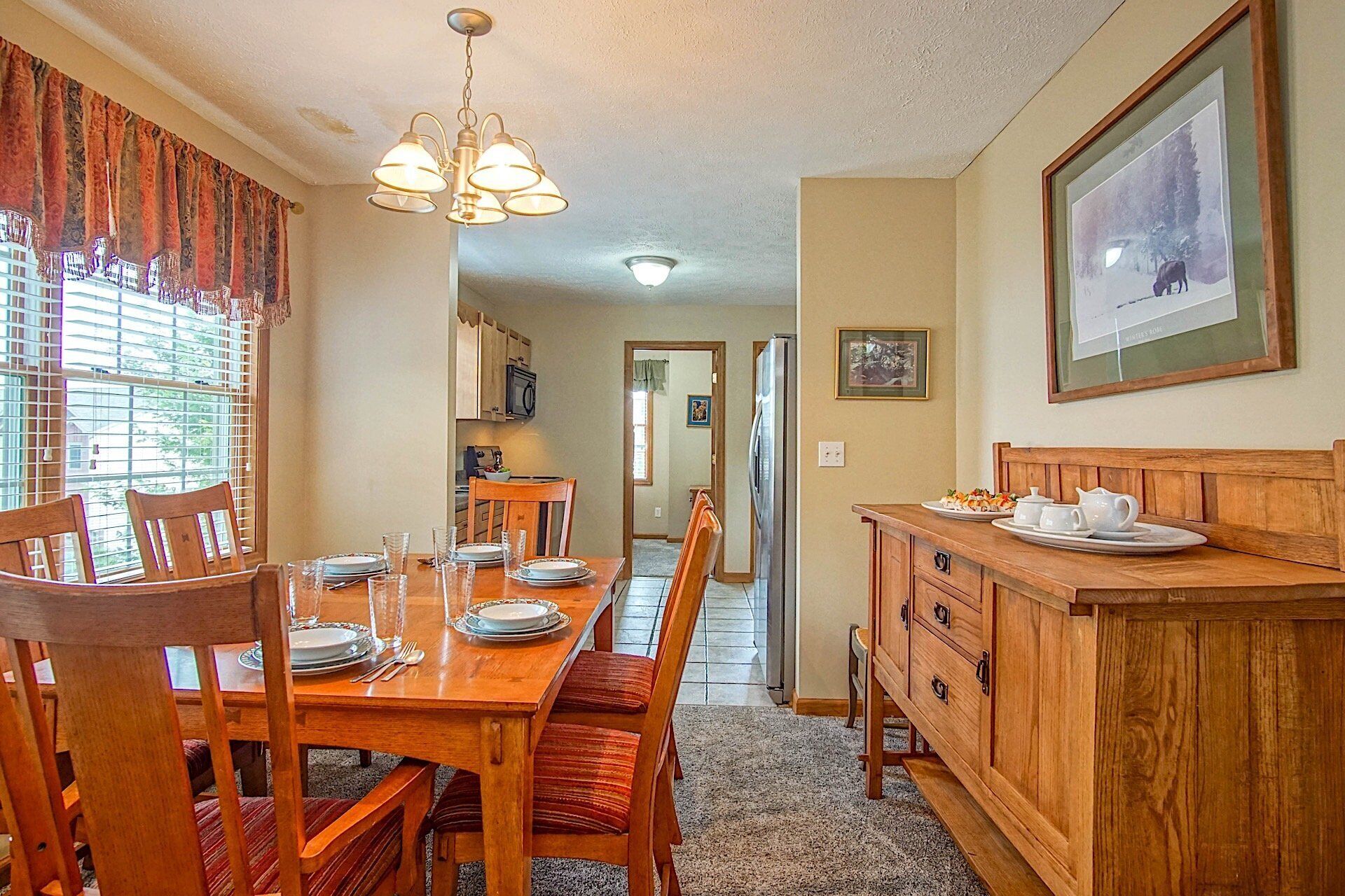 A dining area with a wooden table set for four, a chandelier, and a kitchen visible in the background.