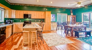 Une cuisine avec des meubles en bois, un îlot central doté de tabourets, un coin repas avec une table et des chaises, et une grande fenêtre.