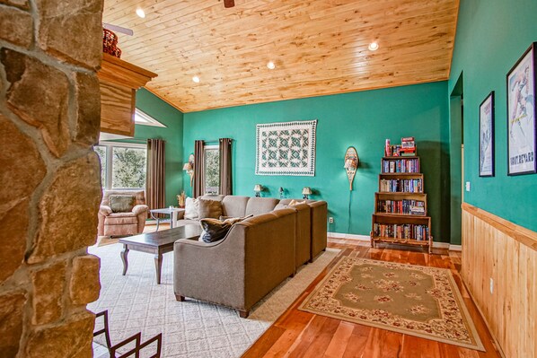A cozy living room with a stone wall, wooden ceiling, and teal walls. There is a gray sofa, a wooden coffee table, and a bookshelf with books. - South West Ridge 72-153 5 Br: Large Western Views. Private hot Tub. Wood Fireplace (Snowshoe)