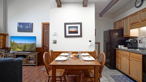 Un comedor con una mesa de madera dispuesta para cuatro, rodeada de sillas de madera. Un televisor está montado en la pared a la izquierda.