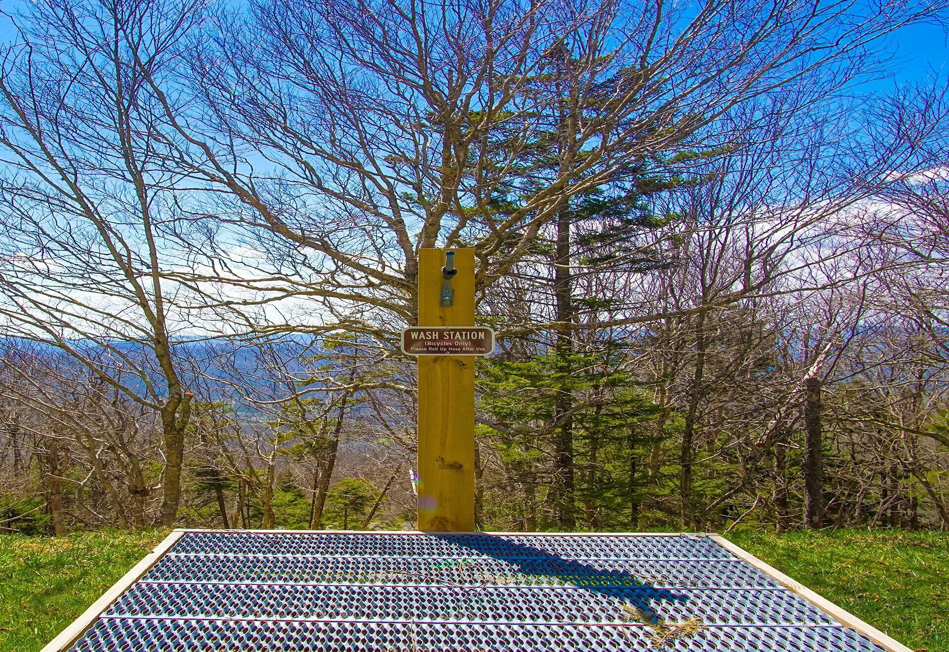 A wash station sign on a wooden post with a metal grate below, surrounded by trees and a clear sky.