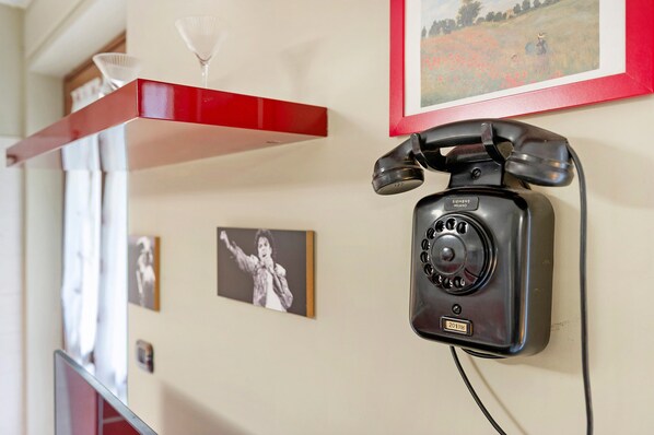Ein altes Telefon an der Wand, umgeben von gerahmten Bildern und einem roten Regal darüber.