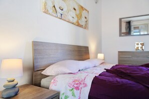 Ein ordentlich bezogenes Bett mit einer violetten Decke, ein Nachttisch aus Holz mit einer Lampe und ein an der Wand befestigter Spiegel.