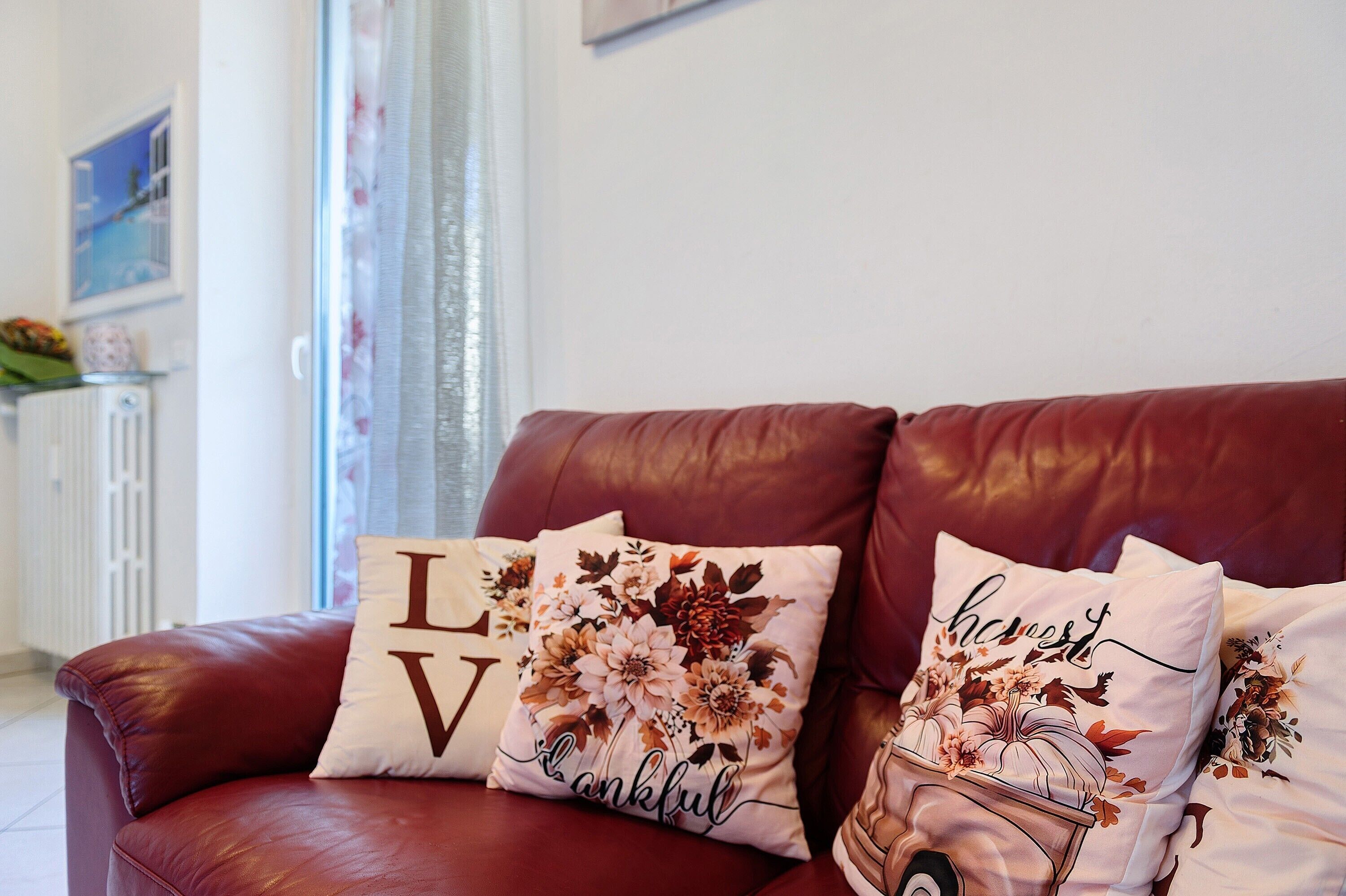A red leather sofa with floral and text pillows, a radiator, and a window with curtains.