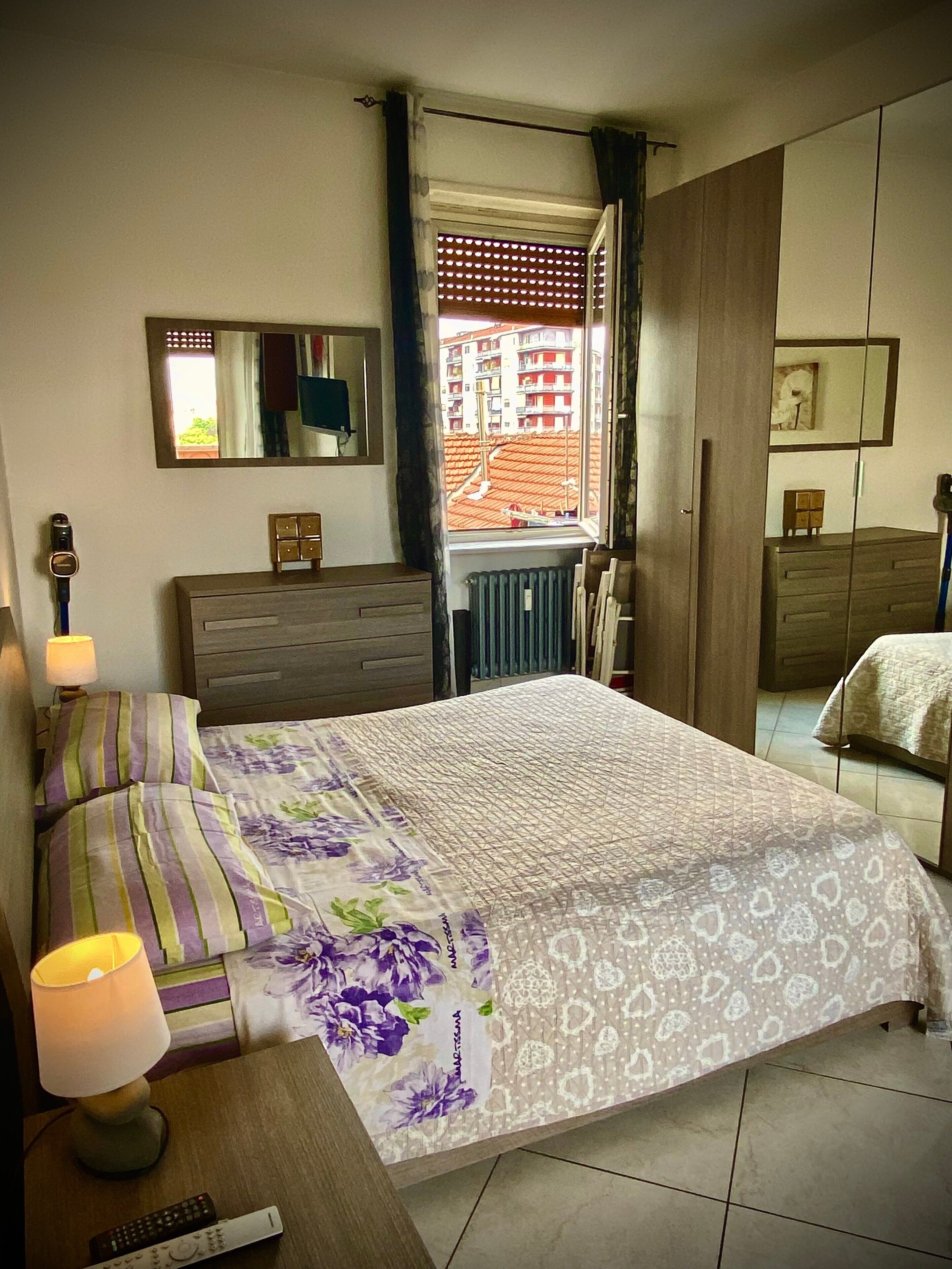 Ein Schlafzimmer mit einem Bett, Nachttischlampen, einem Schminktisch, einem Spiegel und einem Fenster mit Blick auf Gebäude.