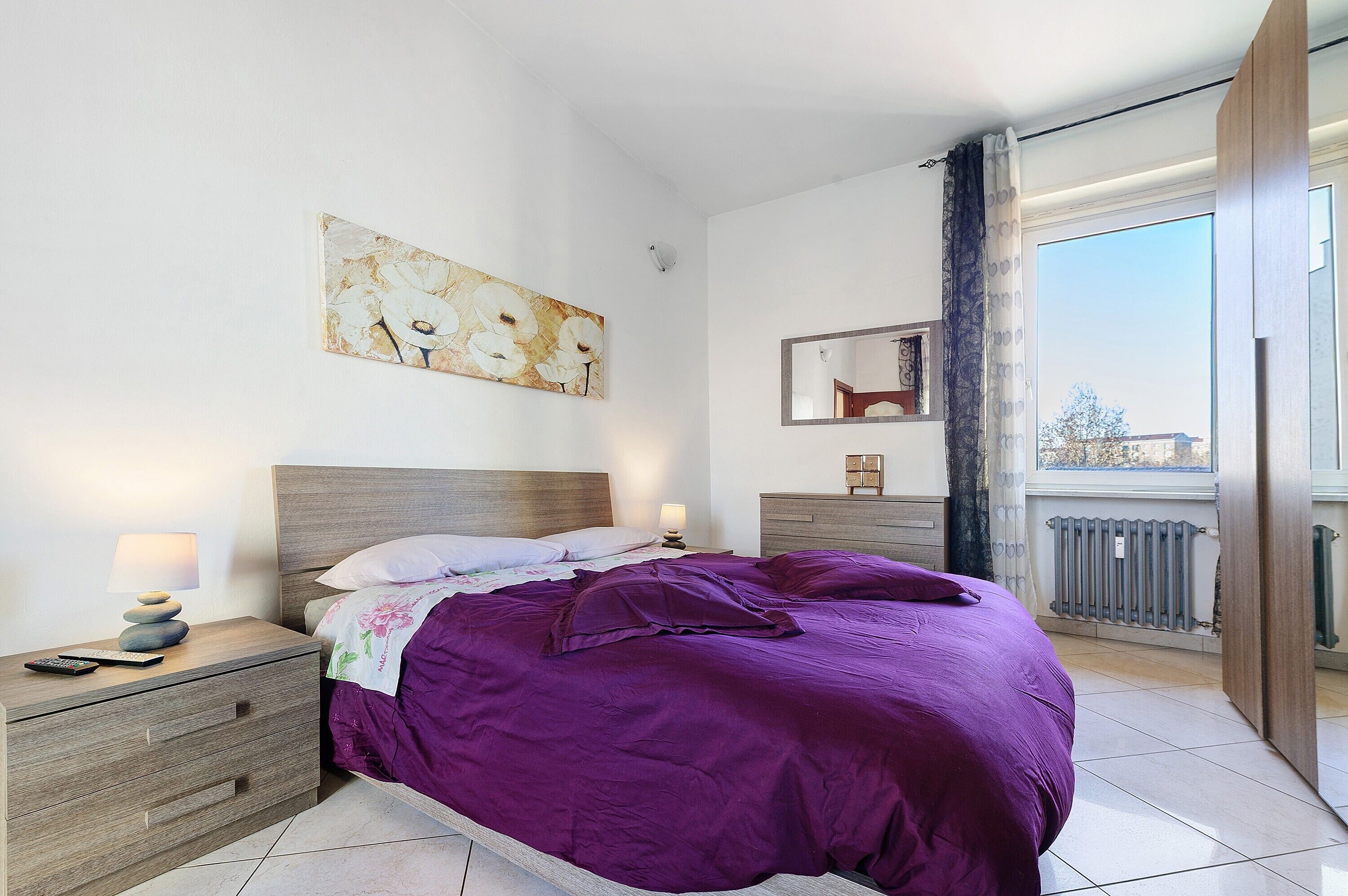 Une chambre à coucher avec un lit, une table de chevet, une lampe et une œuvre d’art accrochée au mur.