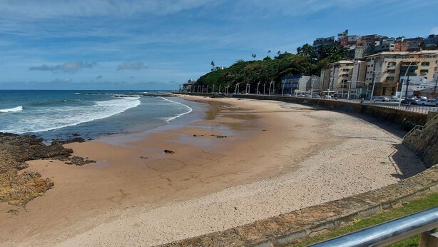 A beach with a rocky shoreline, a paved walkway, and buildings in the background. - COOND0102 - Ondina Residence (Salvador)