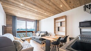 Een moderne woonkamer met een houten plafond, een bank, een eettafel met stoelen, en een keuken met een spoelbak en een magnetron.