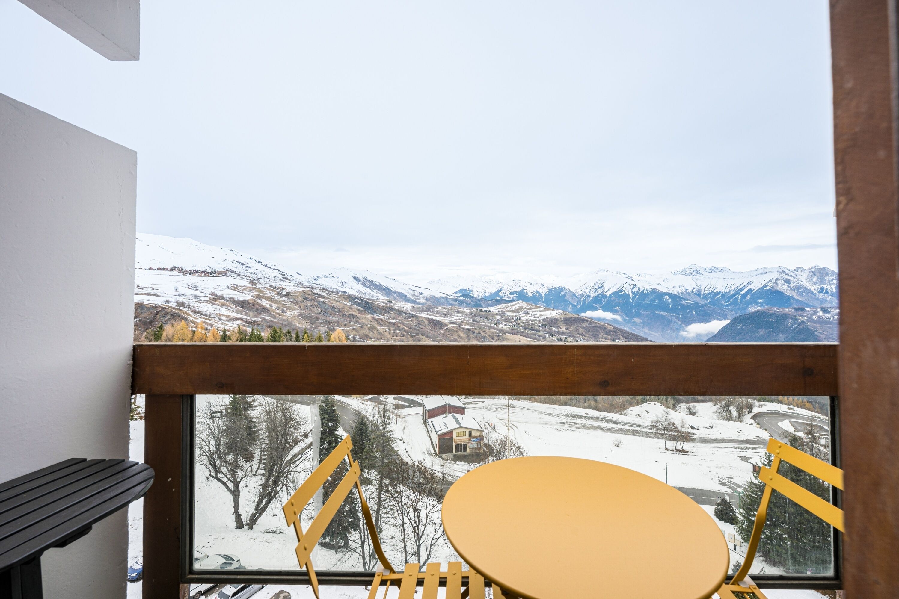 Ein Balkon mit Holzgeländer, einem gelben Tisch und zwei gelben Stühlen, mit Blick auf eine verschneite Berglandschaft, in der Ferne ein kleines Gebäude.