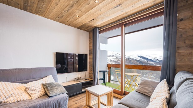 Een moderne woonkamer met een houten plafond, een grijze bank, een houten salontafel en een groot raam met uitzicht op bergen.