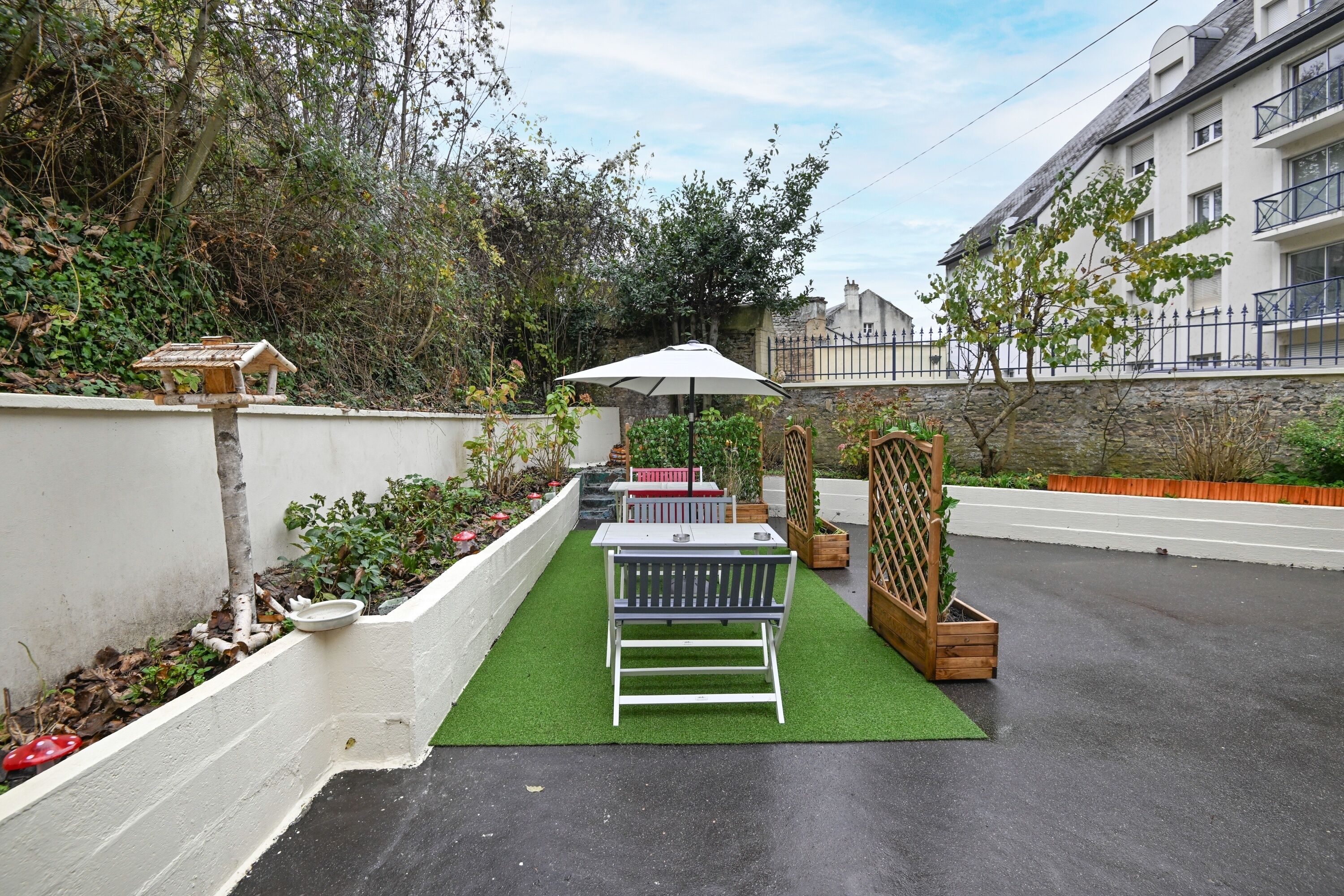 A patio area with a white bench, a table, and an umbrella. There is a small garden with plants and a bird feeder.