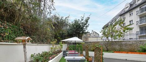 A patio area with a white bench, a table, and an umbrella. There is a small garden with plants and a bird feeder.