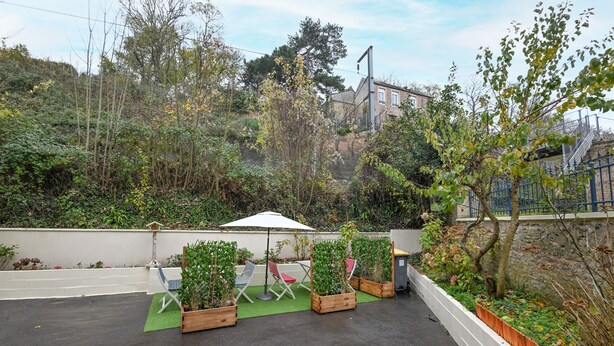 An outdoor patio area with a table and chairs, surrounded by planters and a view of a hill with trees and a building in the background. - Le 4 du Clos Fleuri - Proche Hippodrome (Caen)