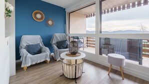A room with a blue wall, two covered chairs, a round table with a woven top, and a large window with a view of the outdoors. - La Résidence Le Belvédère - Avec Balcon (Le Tholy)