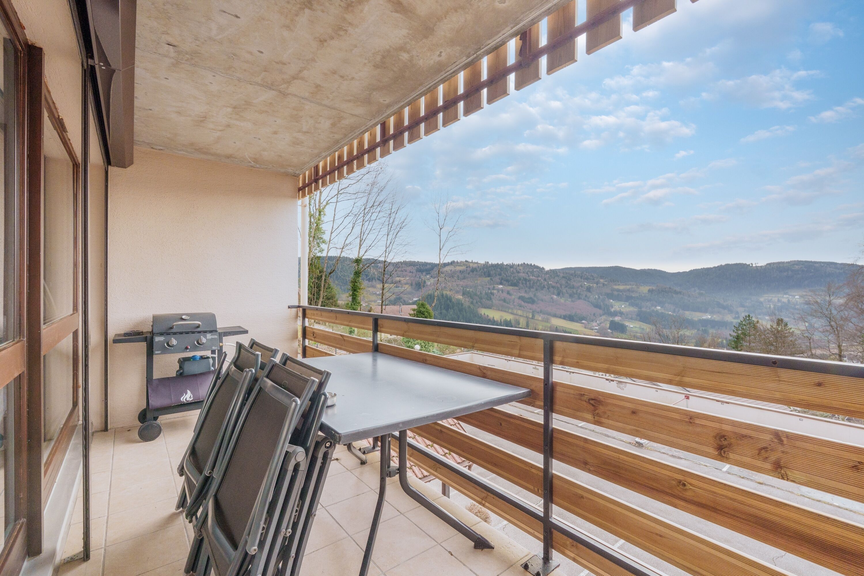 A balcony with a table and chairs, a grill, and a view of a hilly landscape.