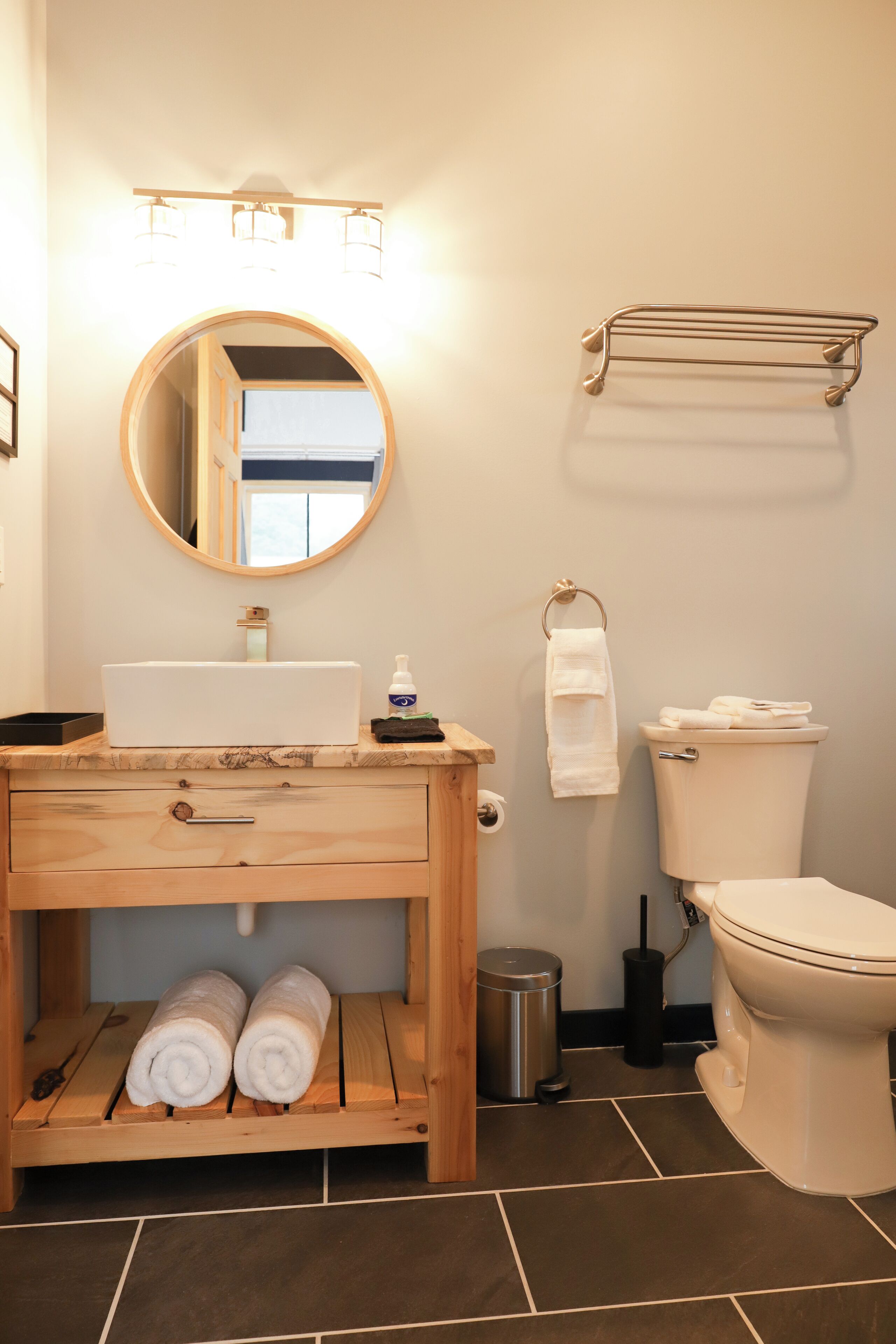 Une salle de bain avec un meuble-lavabo en bois, un miroir rond, des toilettes et un porte-serviettes.