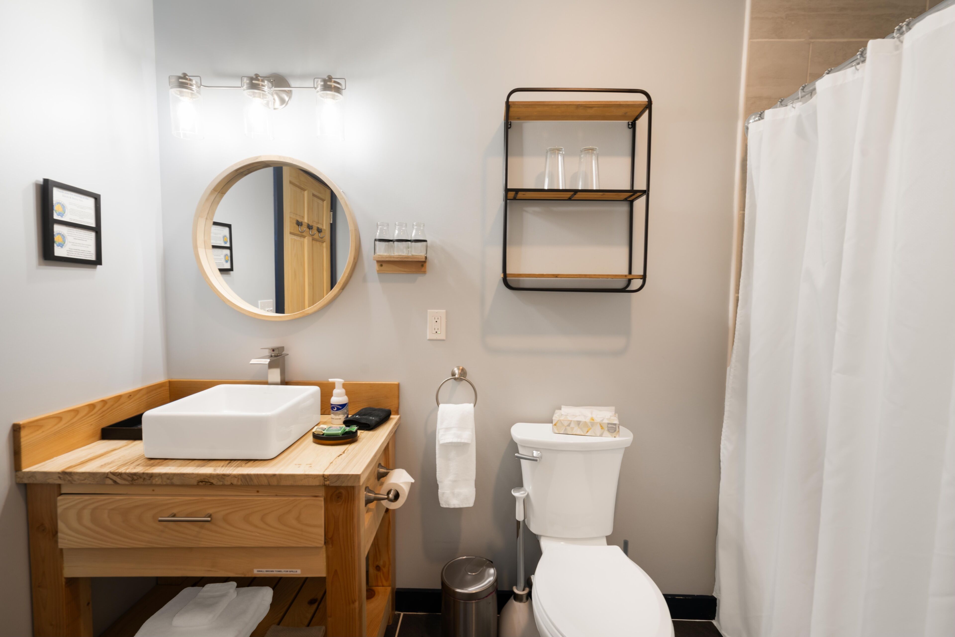 A bathroom with a wooden vanity, a round mirror, a toilet, and a shower curtain.