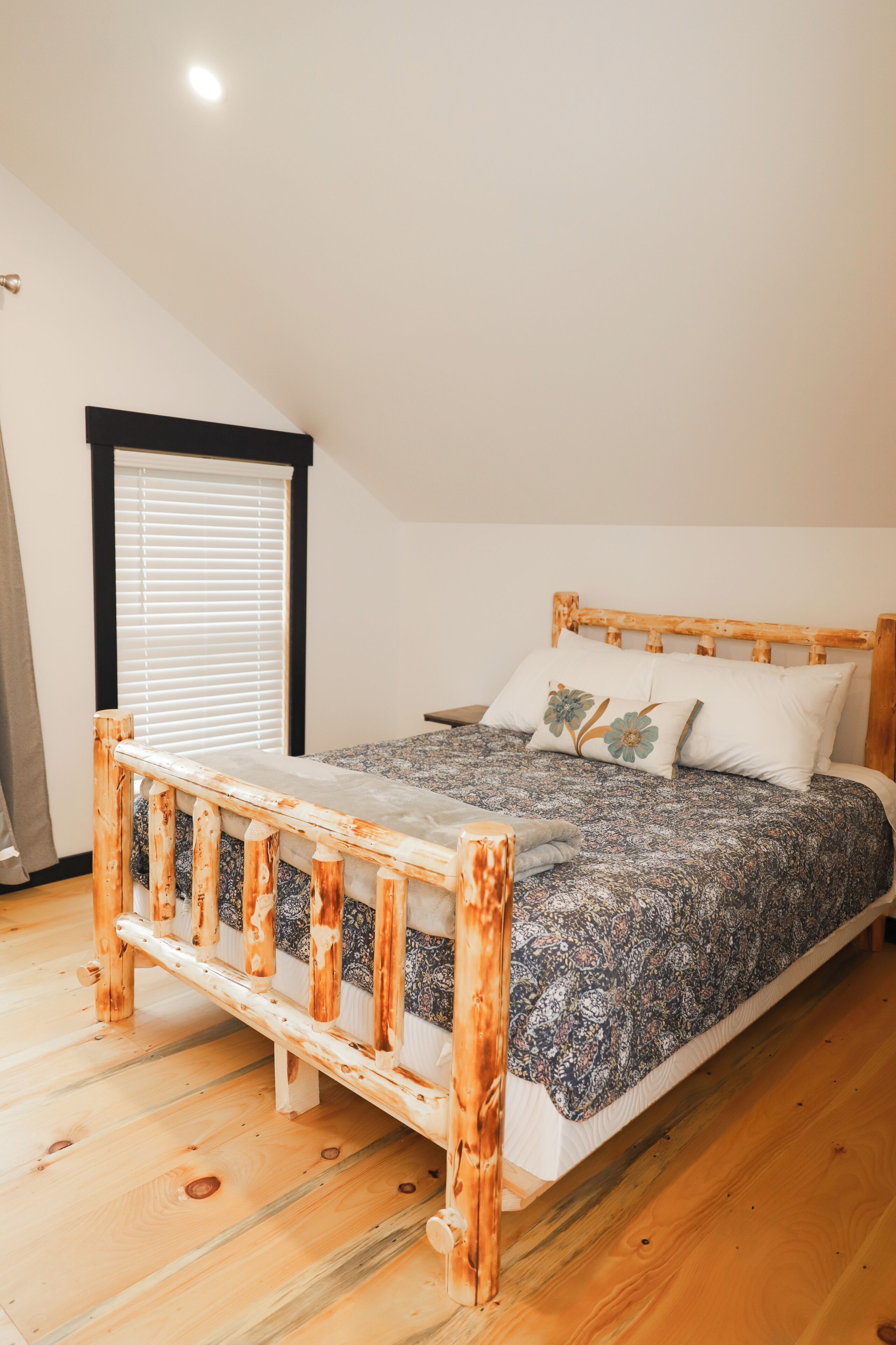 Un dormitorio con estructura de cama de madera, colcha estampada y ventana con persianas.