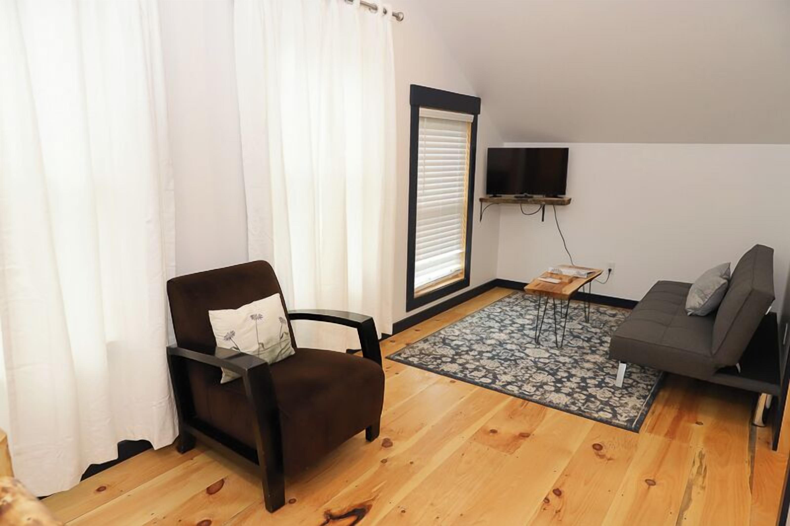 A living room with a brown armchair, a gray sofa, a television mounted on the wall, and a patterned rug on the wooden floor.