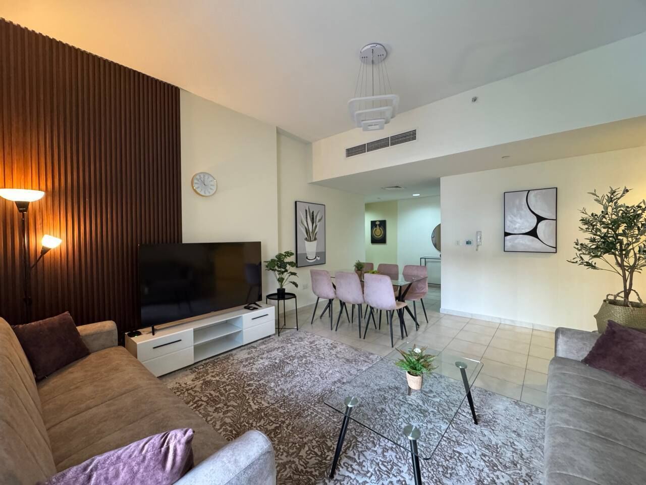 A modern living room with a flat-screen TV, a dining area with pink chairs, and a large potted plant.