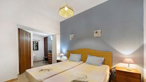 Habitación de hotel con dos camas, cabecera de madera, mesita de noche con lámpara y puerta que da a otra habitación.