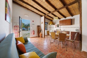 Un salon avec un canapé, des fauteuils, un coin repas comprenant une table et des chaises, une cuisine avec des placards et un sol carrelé.