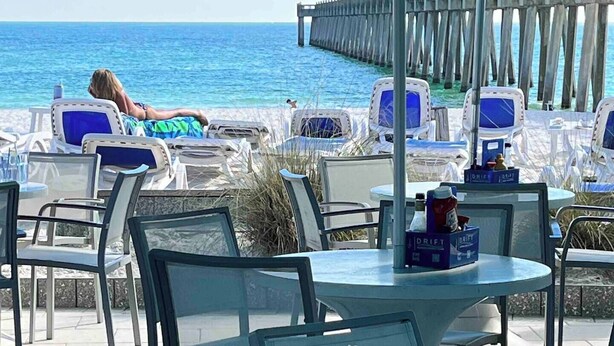 Un patio frente a la playa con mesas y sillas, un muelle de madera al fondo y una persona recostada en una silla de descanso.