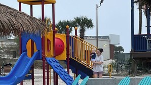 Un parque infantil en la playa con toboganes y una casita, rodeado de arena y palmeras.