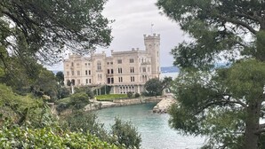 A large, historic building with a tower, situated by a body of water, surrounded by lush greenery. - Double suite with sea view (Trieste)