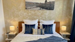 A neatly made bed with a blue blanket, two bedside lamps, and a framed cityscape picture on the wall. - Double suite with sea view (Trieste)
