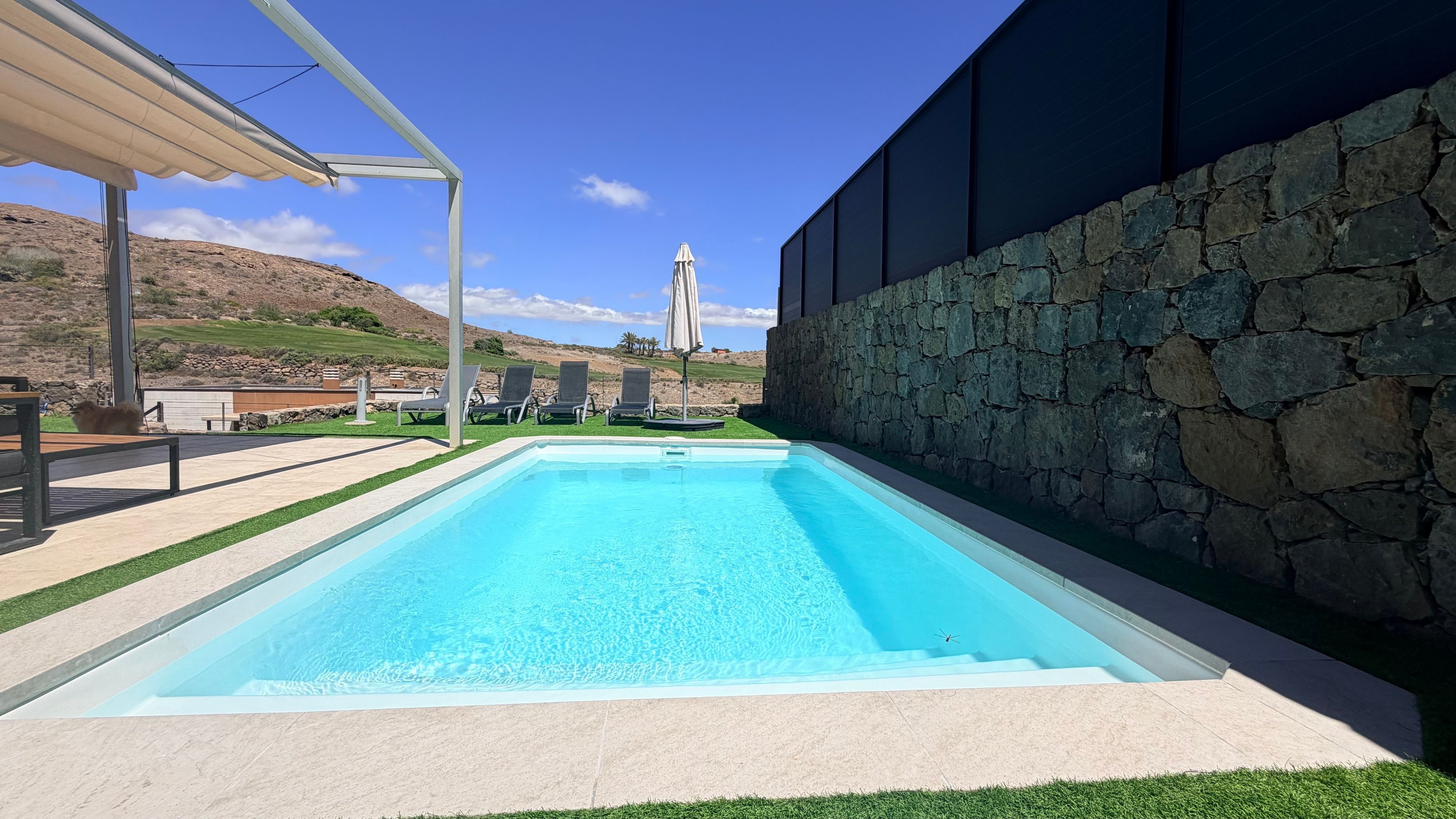 Une piscine rectangulaire, avec un mur en pierre d’un côté et un espace abrité, équipé de chaises et d’un parasol, de l’autre.