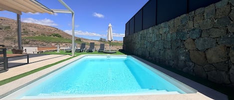A rectangular swimming pool with a stone wall on one side and a covered area with chairs and an umbrella on the other.