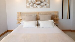 Une chambre avec une tête de lit en lattes de bois, deux oreillers beige, un couvre-lit blanc, une grande œuvre d’art florale et un miroir sur le mur.