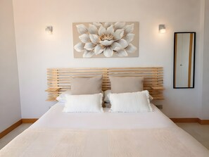 Camera da letto con testiera in legno a listelli, due cuscini beige, copripiumino bianco, un’ampia opera d’arte floreale e uno specchio a parete.