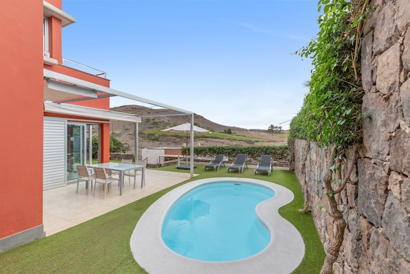 Una piscina a forma curva, circondata da un patio con tavolo e sedie, e da una zona coperta con poltrone relax.