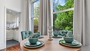 Une table de salle à manger ronde, entourée de chaises turquoise et agrémentée d’assiettes assorties, est placée dans une pièce dotée de grandes fenêtres offrant une vue sur la verdure environnante.