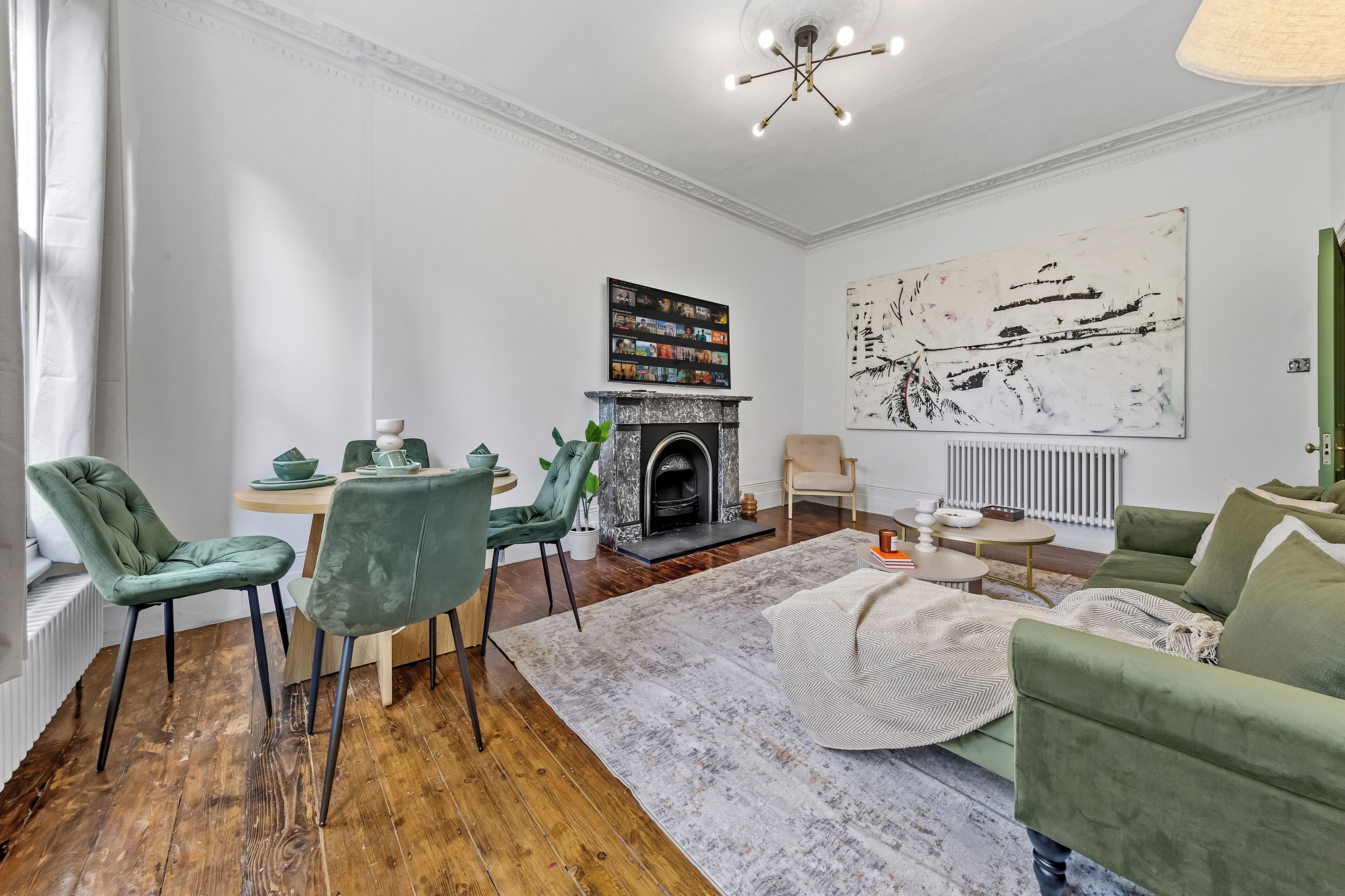 A modern living room with a fireplace, green upholstered chairs, a round coffee table, and a large abstract painting on the wall.