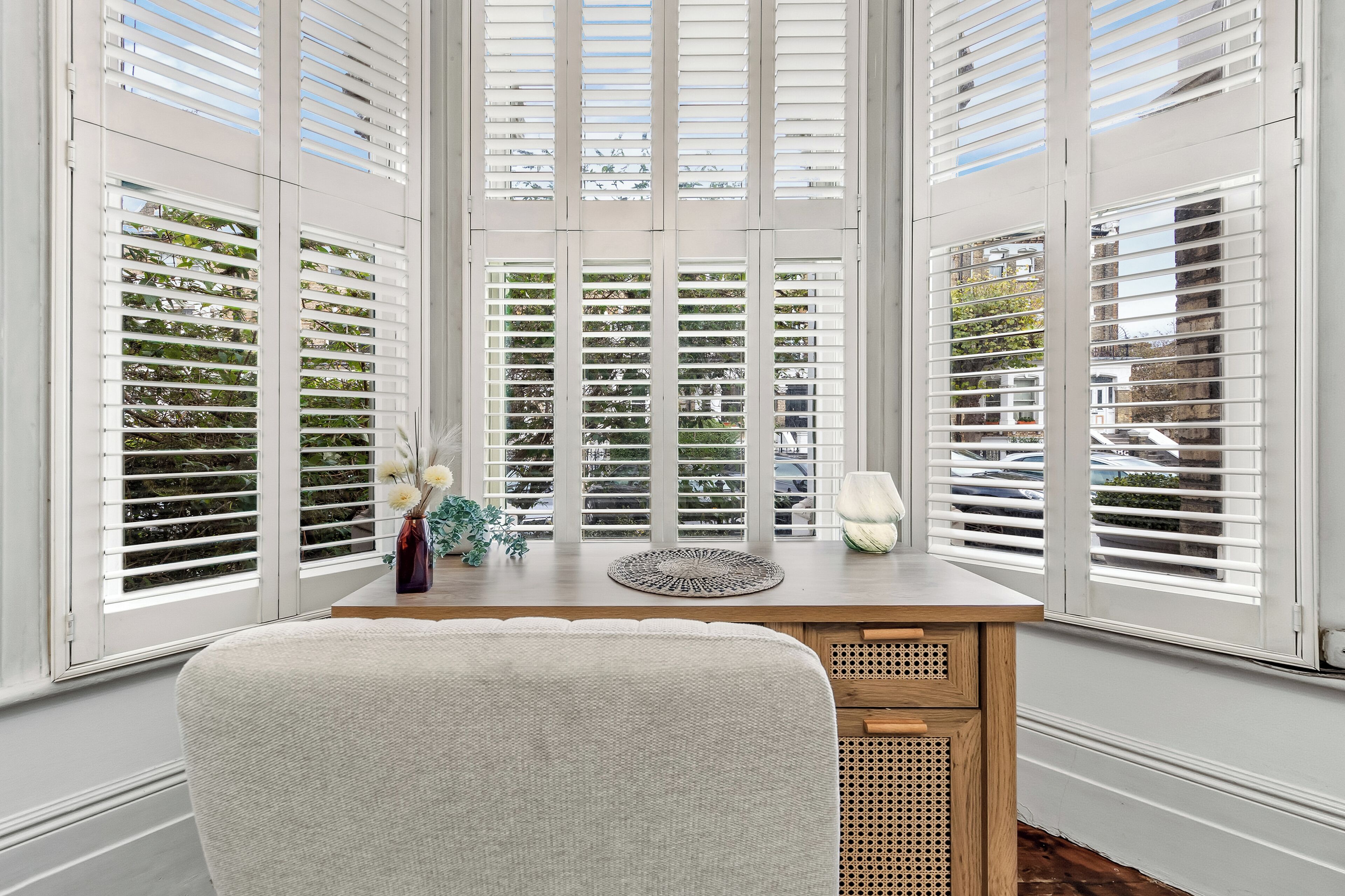 Una habitación con un escritorio de madera, contraventanas blancas y vistas a coches aparcados fuera.