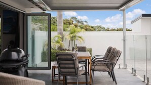 An outdoor patio with a table and chairs, a glass door leading to a garden, and a clear view of the sky and greenery. - ⭐️ The Wine Country Retreat by Homello ⭐️ | Pool • Wineries • Wedding Venues (Kumeū)