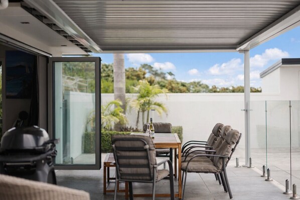 An outdoor patio with a table and chairs, a glass door leading to a garden, and a clear view of the sky and greenery.