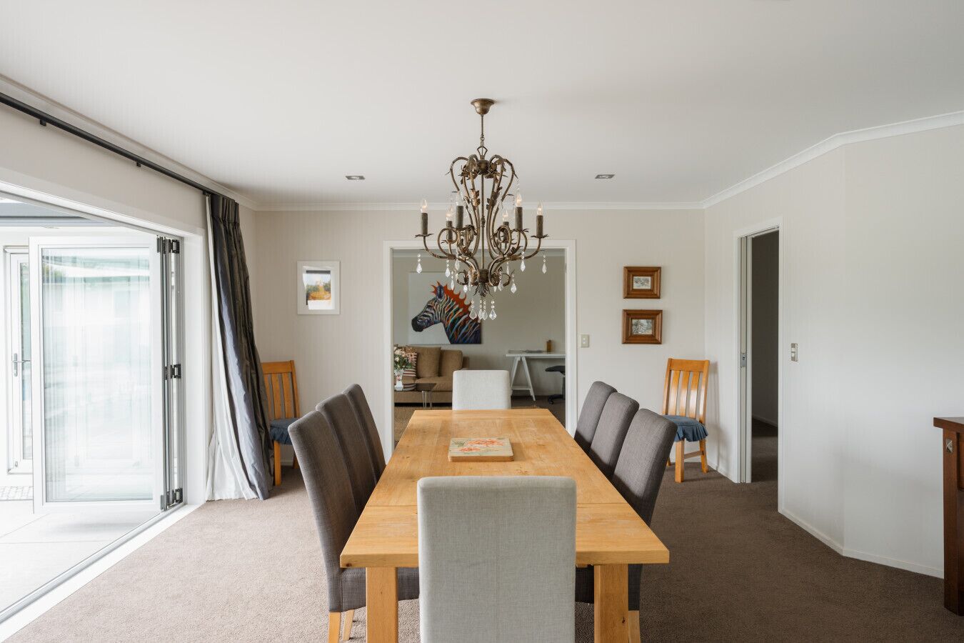 A dining room with a wooden table, grey upholstered chairs, a chandelier, and a large window with curtains.