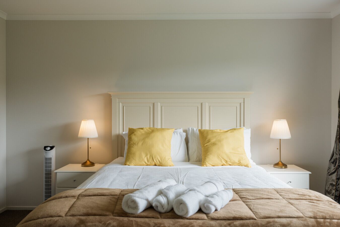 A neatly made bed with white and yellow pillows, a brown quilt, and rolled towels on a white nightstand.