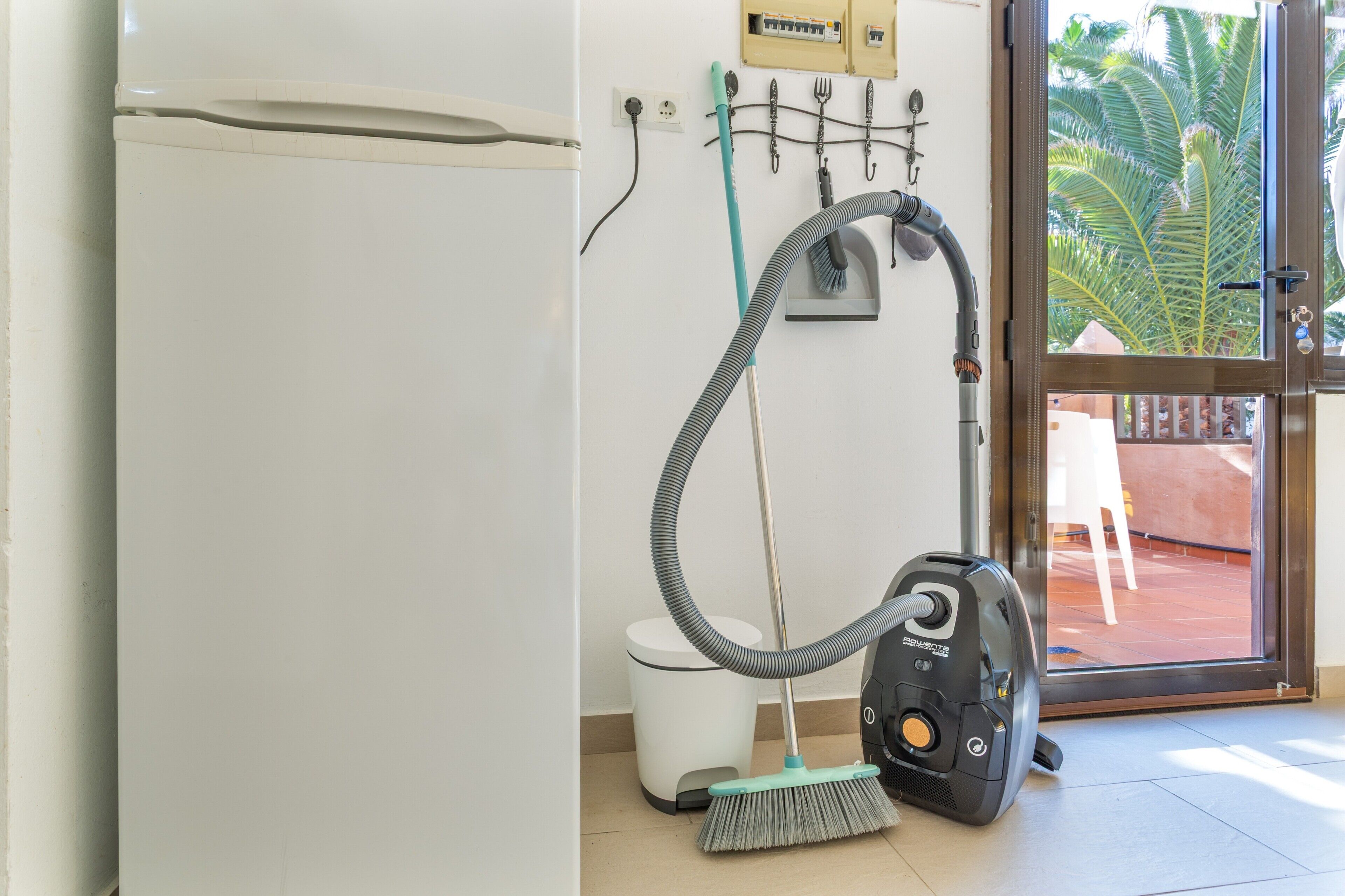 A bathroom with a toilet, a vacuum cleaner, and a window with a view of palm trees.