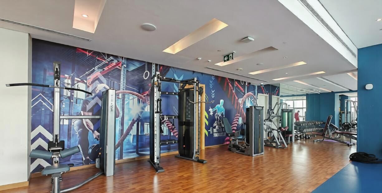 Une salle de sport équipée de divers appareils d’exercice, dotée d’un plancher en bois et d’un mur bleu orné d’une fresque.